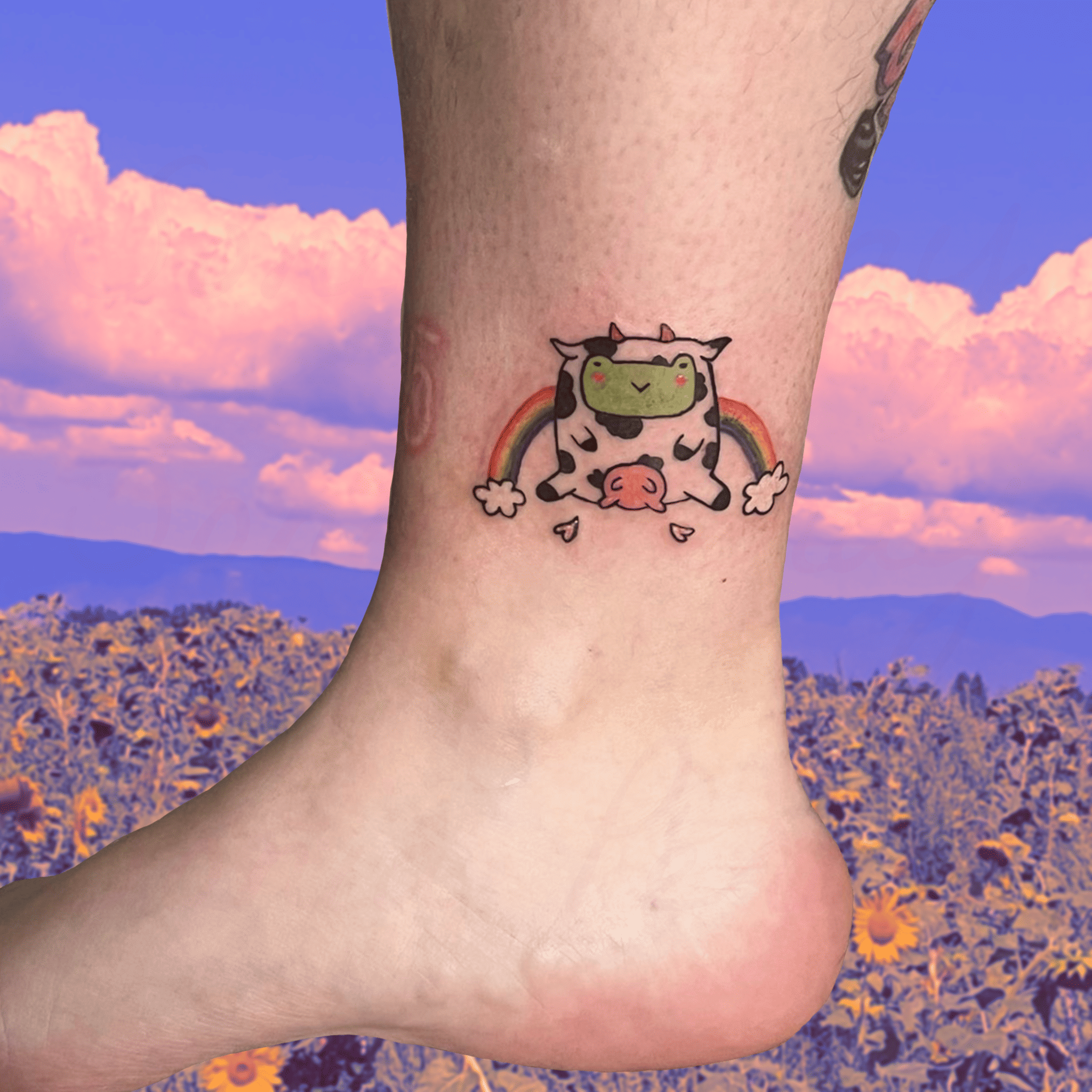 Tatouage en couleur et en freehand d'une grenouille dans un costume de vache avec un arc en ciel derrière
